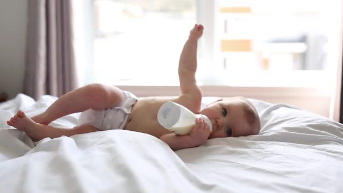 漂亮的女婴躺在床上从瓶子里喝水。儿童尿布