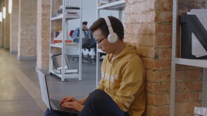 戴着耳机的亚洲潮人在走廊地板上的笔记本电脑上工作
