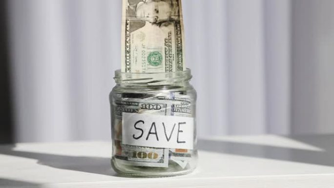 男性手将20美元放在一个玻璃罐中，上面刻有save字样。省钱的概念。