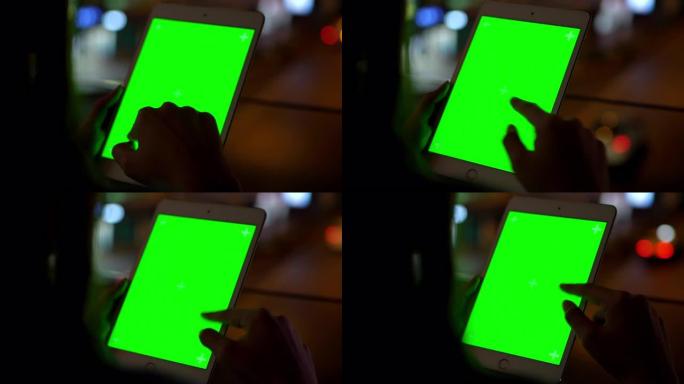 4k近距离后视图亚洲女性的手拿着绿屏数字平板电脑，并在夜间背景下用模糊的城市街道刮擦触摸屏。具有技术