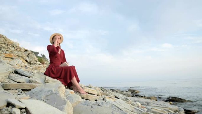 一个迷人的高加索女孩的肖像，穿着一件夏天的红色连衣裙，戴着一顶草帽，怀里抱着一本书坐在海边的石头上，