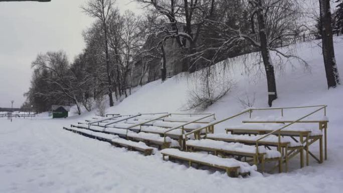 白雪皑皑的长椅在冬天的夏季海滩上，白天在河边。季。白雪覆盖的观众看台