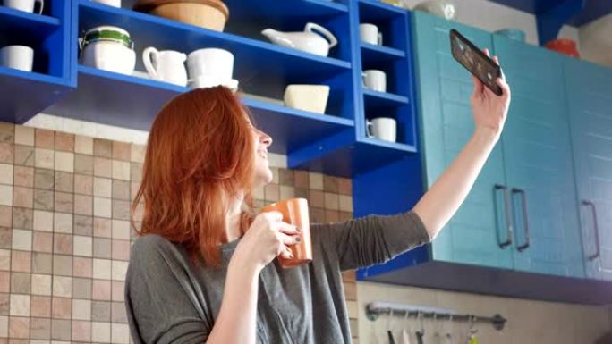 红头发的迷人女孩在家庭厨房喝早间咖啡。在社交网络的智能手机上自拍。在现代厨房用手机自拍的漂亮女人。