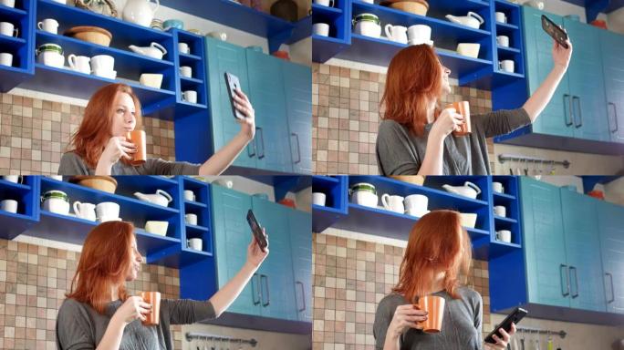 红头发的迷人女孩在家庭厨房喝早间咖啡。在社交网络的智能手机上自拍。在现代厨房用手机自拍的漂亮女人。