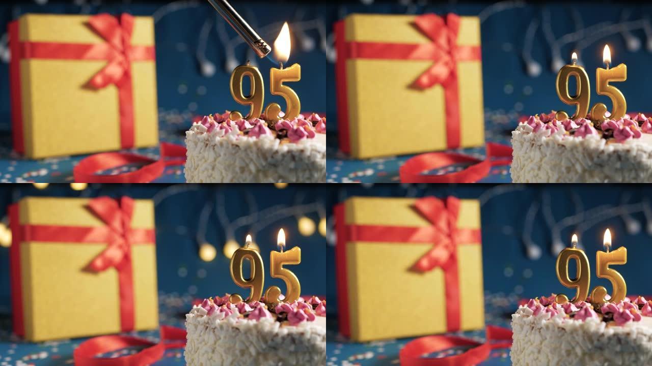 白色生日蛋糕95号金色蜡烛被打火机燃烧，蓝色背景带灯和礼物黄色盒子用红丝带绑起来。特写