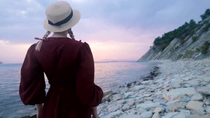 从后面看。一个穿着红色连衣裙和草帽的年轻女孩的肖像在海边的岩石海岸和粉红色的日落。等待水手们的归来