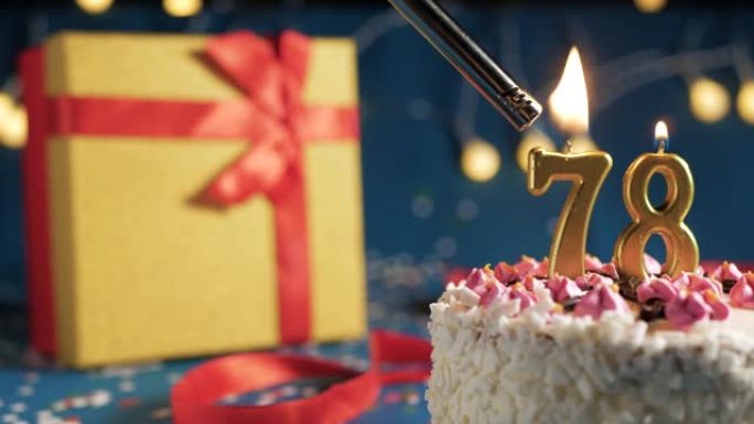 白色生日蛋糕编号78点灯燃烧的金色蜡烛，蓝色背景带灯和用红丝带绑起来的礼物黄色盒子。特写
