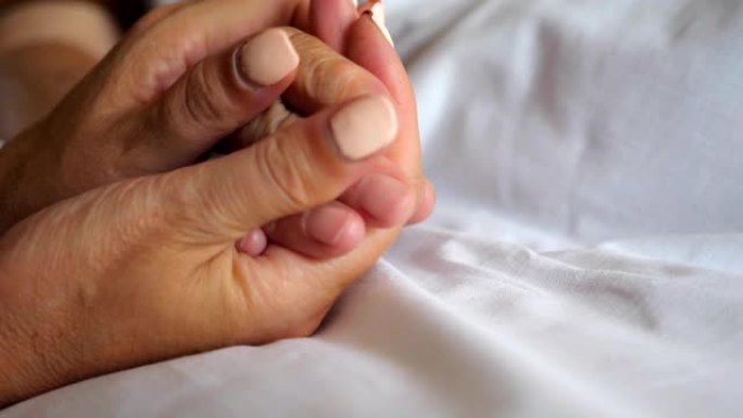 女人牵着老母亲的手躺在床上，表现出关怀或爱心。女儿在医院安慰年迈的妈妈皱巴巴的手臂。孙女温柔地抚摸奶