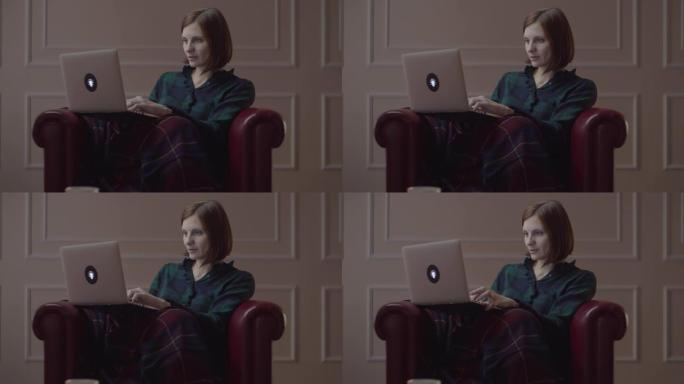 30多岁的年轻工作女性坐在舒适的扶手椅上，用格子在笔记本电脑上打字。女性独自在家工作。