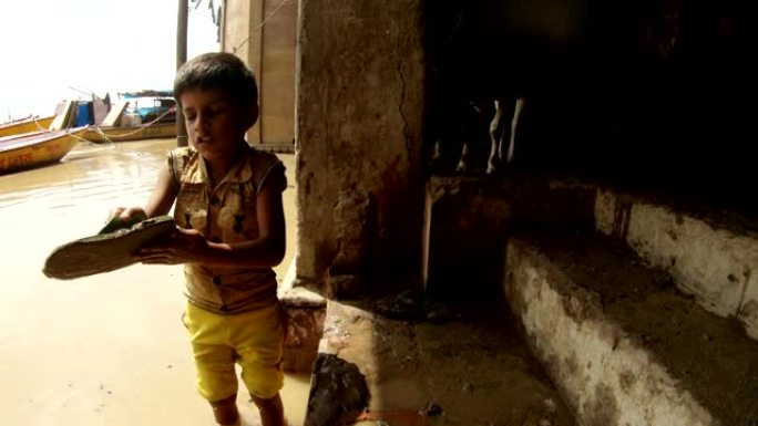 小印度男孩用手清除他的拖鞋从泥水淹没的圣河恒河肮脏的水牛和船在码头的背景瓦拉纳西
