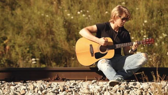 精力充沛的年轻人戴着墨镜坐在铁路上，用原声吉他表演