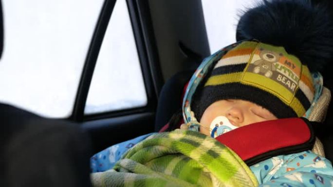 小孩睡在移动的汽车里。孩子系好安全带，冬天在外面，他穿着暖和的衣服。阳光照在他的眼睛里。特写