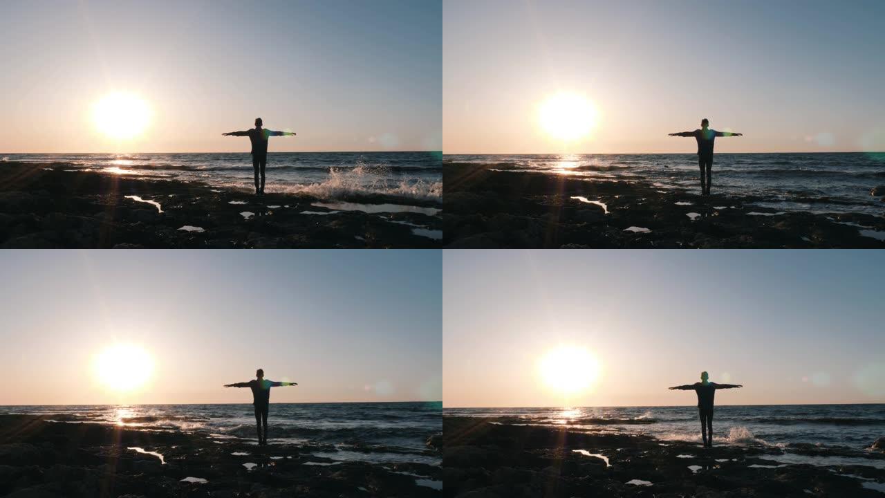 年轻人站在海边的剪影，双手举起。苗条的男人站在岩石海滩上，面对美丽的日落。快乐的人举起双手，看着地平