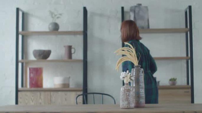 30多岁的年轻女性从架子上拿花瓶和花盆，放在木桌上。简约室内公寓。女人欣赏室内物品。