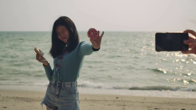 享受生活的女性朋友吃甜甜圈甜美味拍照在海滩放松和自由，迷人的亚洲女性旅行者的生活方式。4K UHD镜