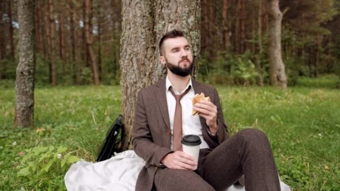 年轻迷人的男性商人穿着棕色西装，公文包坐在公园树下的草地上，休息，吃三明治和喝咖啡。