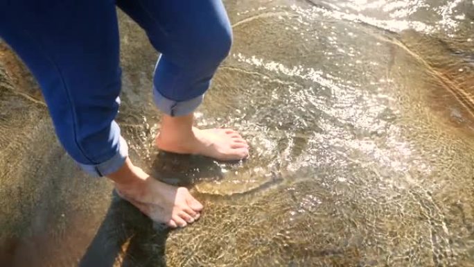 赤脚的年轻女子在缓慢流动的溪流中的水中飞溅和踢腿。女士们在瀑布享受大自然的假期。暑假生活方式的概念。