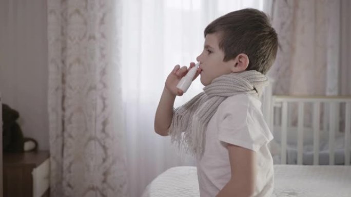 穿着温暖围巾鼻塞的生病的男孩在流感期间坐在私人公寓的床上使用疾病滴剂