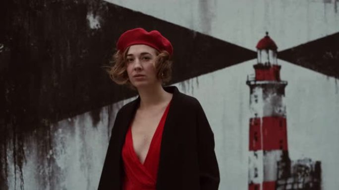 穿着外套和红色连衣裙的年轻女子站在墙壁附近，并带有灯塔的形象。短卷发的漂亮女人。