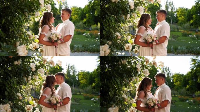 新郎和新娘微笑着亲吻茂密的白玫瑰丛。新婚夫妇互相拥抱，深情地望着对方的眼睛。一对穿着白色婚纱的漂亮夫