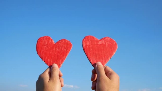 双手握着蓝色天空背景上的两个红色心形回收纸板。情人节的爱情象征。爱情和浪漫的概念。
