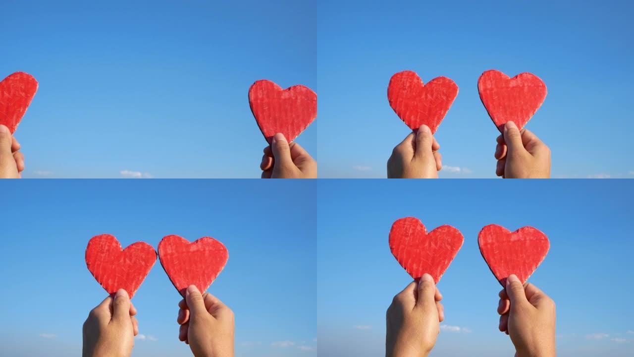 双手握着蓝色天空背景上的两个红色心形回收纸板。情人节的爱情象征。爱情和浪漫的概念。