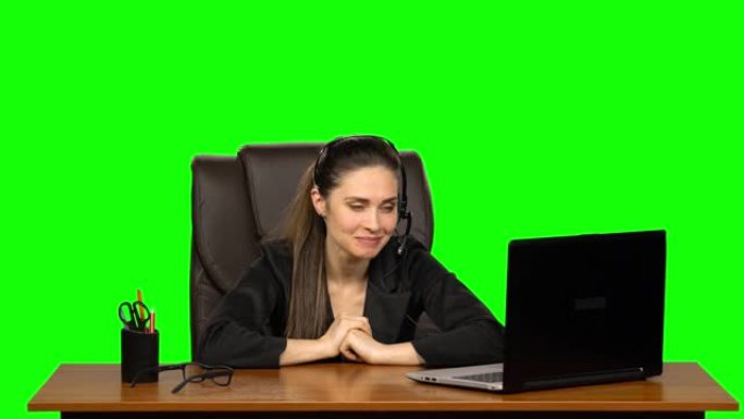 戴着耳机的女商人用网络摄像头说话，带着喜悦的心情看着笔记本电脑。绿屏