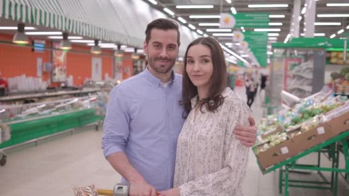 迷人的快乐年轻千禧一代家庭夫妇用购物车在超市购物，看着相机微笑，可爱的夫妻买家顾客满意杂货店概念