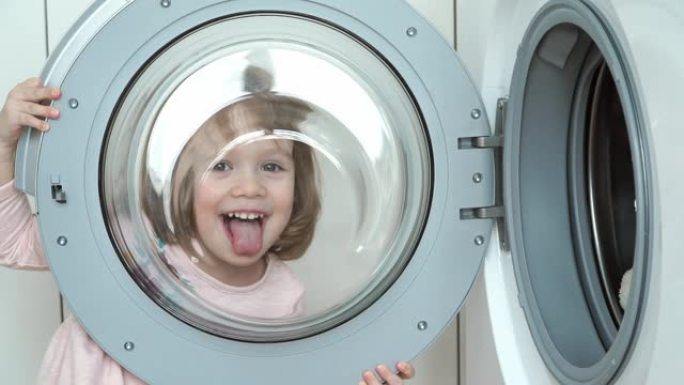 小滑稽女孩玩洗衣机舱口的特写。孩子透过舱口，周围的傻瓜，微笑和大笑