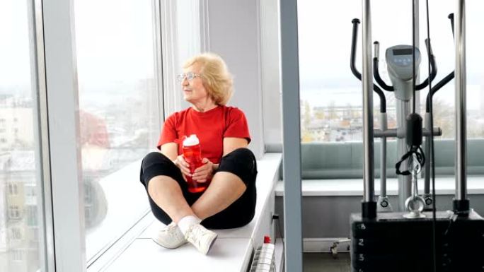 穿着红色t恤的相当活跃的退休妇女坐在健身房的窗台上。老年人拿着瓶子喝水。平静的老奶奶看着健身俱乐部的