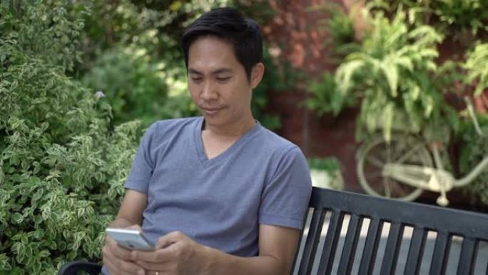 亚洲男子坐在长凳上，在花园里使用智能手机。手持镜头