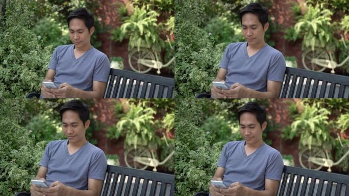 亚洲男子坐在长凳上，在花园里使用智能手机。手持镜头