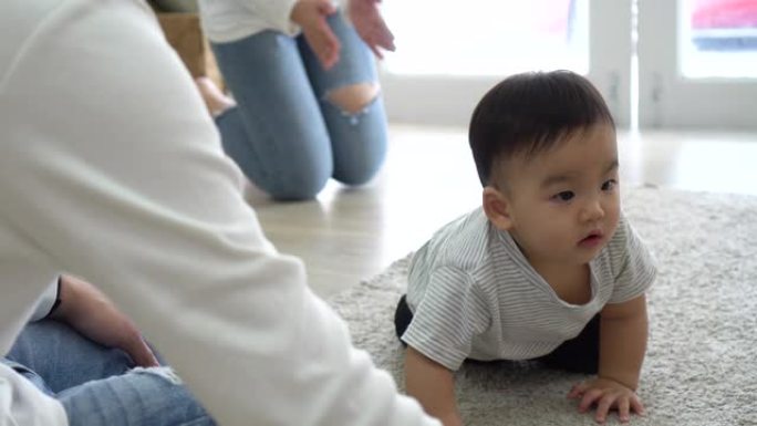 从可爱的亚洲男婴的上方看着妈妈手中的玩具，在地毯上爬向父母，而家人则在家里共度时光