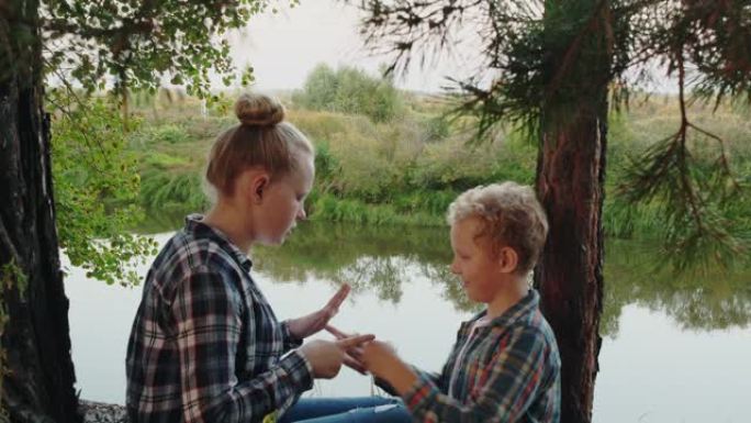 十几岁的女孩和男孩在松树林的河岸上玩石头剪刀。快乐的姐姐和哥哥在河边的夏季森林里玩石头剪刀。