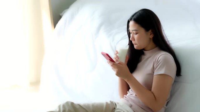 美丽的年轻亚洲女性坐在床边，在阳光下用手机在卧室里网上购物
