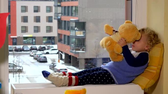可爱的小女孩躺在窗户旁边的散热器上，带着泰迪熊。冬季降雪