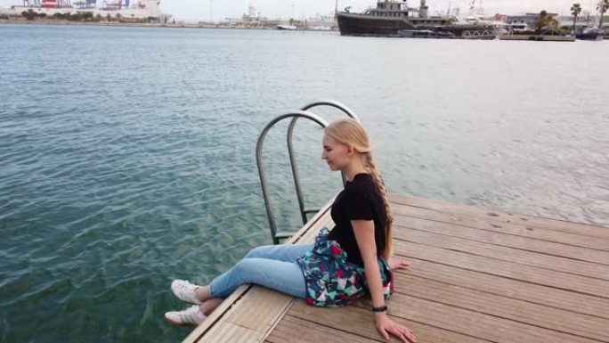 女孩独自坐在港口码头的边缘，停在停着的帆船前，以美丽的景色放松
