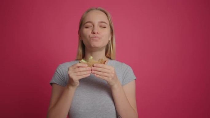 年轻的金发女孩在粉色背景下愉快地吃美味的三明治