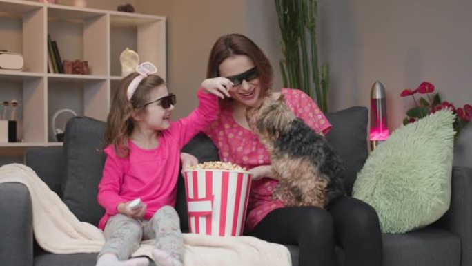 快乐的白人妇女和她戴着3D眼镜的小女儿坐在沙发上，一边吃爆米花一边看电视上的喜剧电影，一边笑
