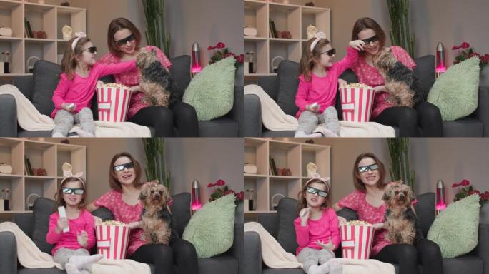 快乐的白人妇女和她戴着3D眼镜的小女儿坐在沙发上，一边吃爆米花一边看电视上的喜剧电影，一边笑