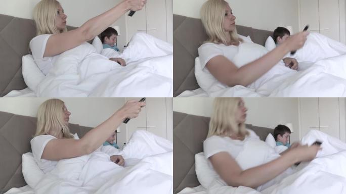 一个金发碧眼的女人躺在床上，通过遥控器切换频道，她的儿子在后台用智能手机玩游戏