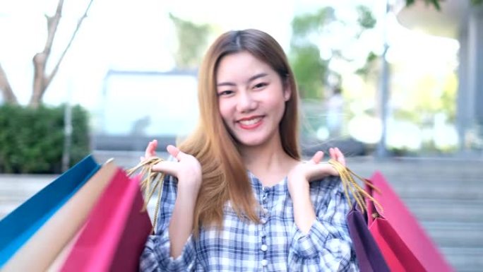 微笑的年轻亚洲女人，购物中心背景上的彩色包。使用智能手机在线购物，站在商场大楼时微笑。生活方式概念