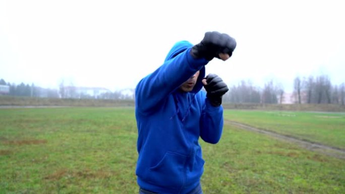 男子运动员拳击手男子戴着拳击手套在户外训练，下蹲和拳打，戴着兜帽的男子看起来咄咄逼人，试图挑衅某人打