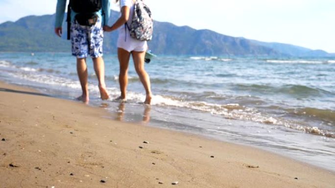年轻夫妇在海洋背景下沿着海滩走。一对带着背包的旅行者在海边行走，泼水洗脚。男人和女人在海浪的沙滩上漫