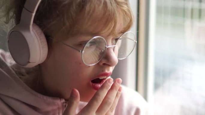 穿着粉色耳机和眼镜的少女坐在家里靠窗的时候无聊。女孩用手打哈欠捂着嘴