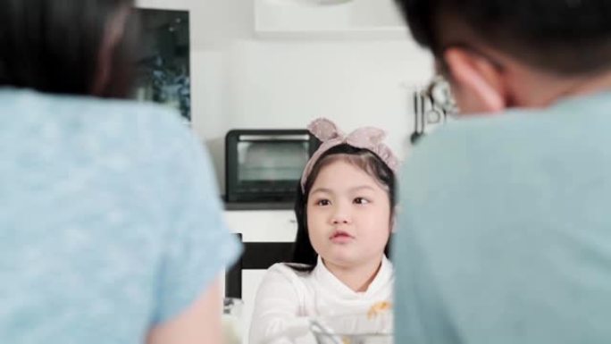 亚洲女孩子无聊的食物和爸爸妈妈试图说服她在餐桌上吃更多的早餐家庭厨房背景