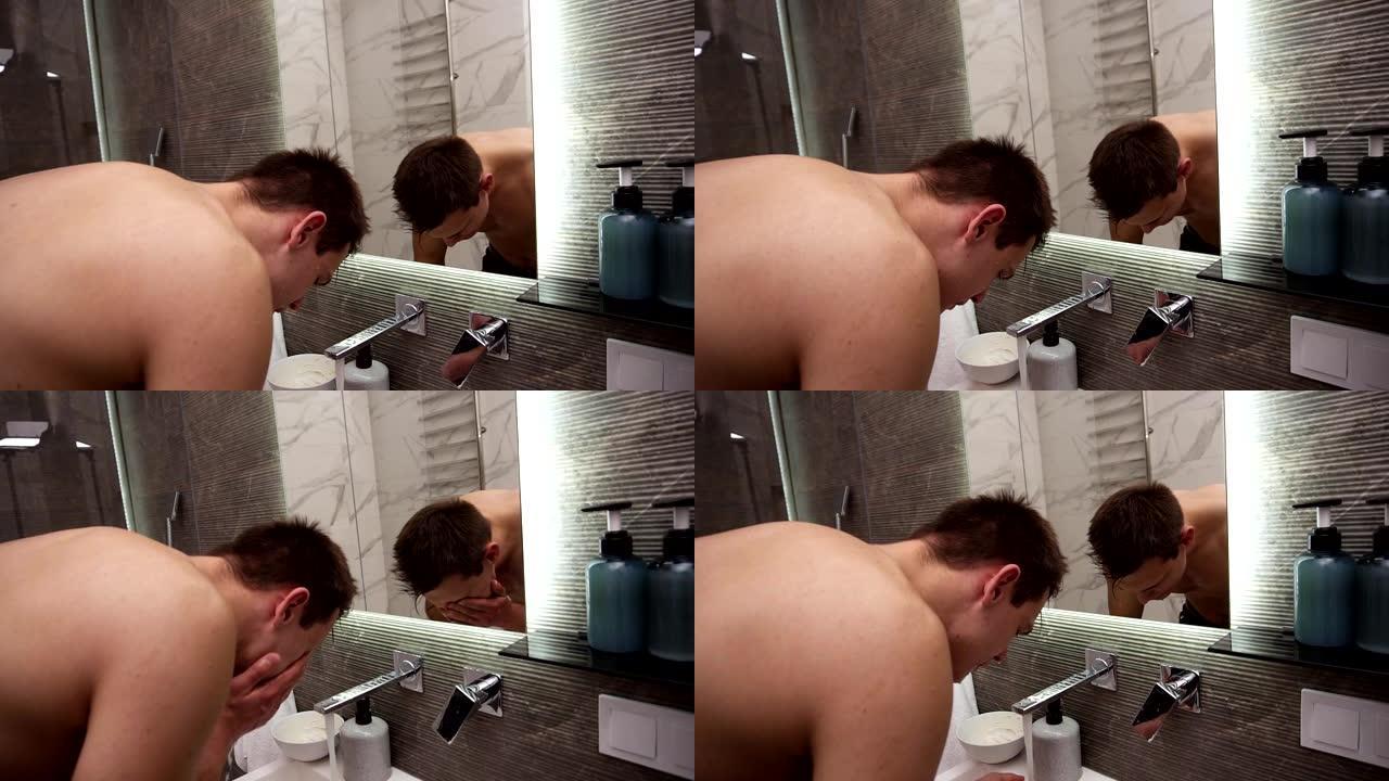 赤裸上身的帅哥在水龙头的冷水下在水槽里洗脸。做早上例行清洁程序的人。看着镜子反射