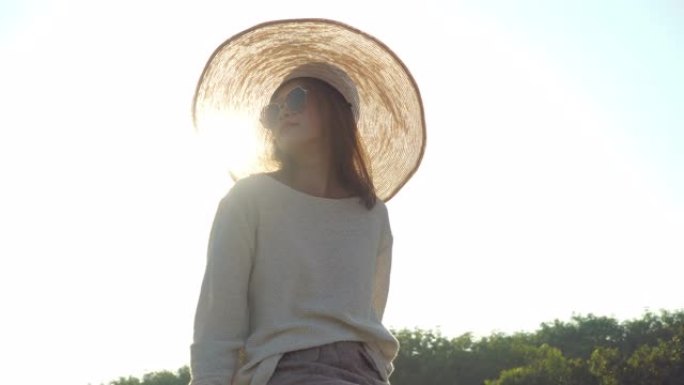 4k年轻美丽快乐的亚洲女人，戴着帽子和太阳镜，肖像微笑着，在夏日日落时享受阳光。快乐的亚洲女人在大自