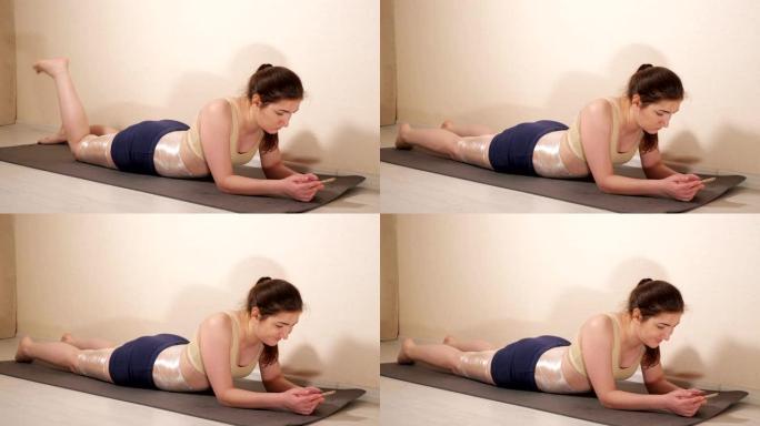 一个女人拿着电话躺在健身垫上。用保鲜膜包裹大腿和腹部