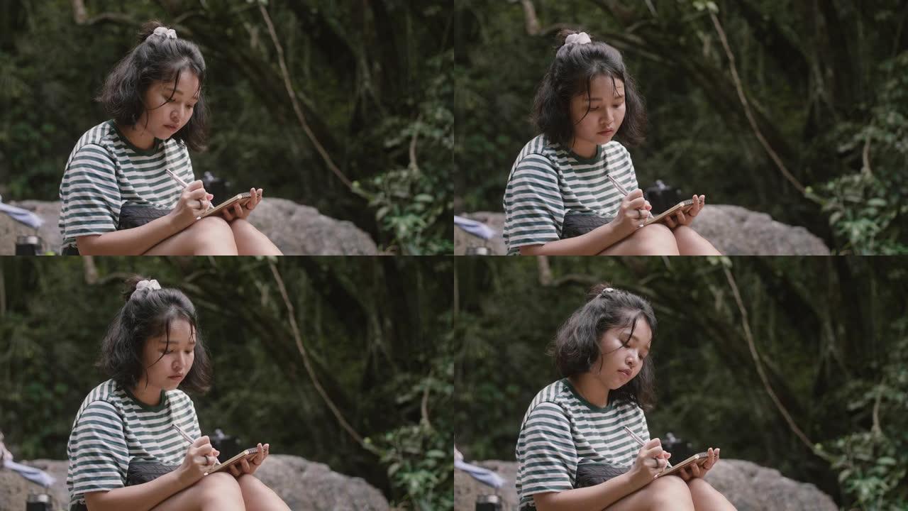 年轻女子坐在瀑布边，在笔记本上画素描。